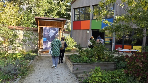 Im Garten der Begegnungsstätte Madeleine Delbrel betrachten zwei Personen ein großformatiges Foto von Madeleine Delbrel. Im Vordergrund sieht man Hochbeete, im Hintergrund ein farbig gestaltetes Haus.