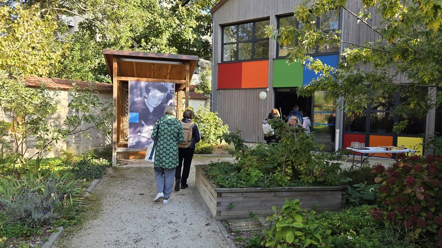 Im Garten der Begegnungsstätte Madeleine Delbrel betrachten zwei Personen ein großformatiges Foto von Madeleine Delbrel. Im Vordergrund sieht man Hochbeete, im Hintergrund ein farbig gestaltetes Haus.
